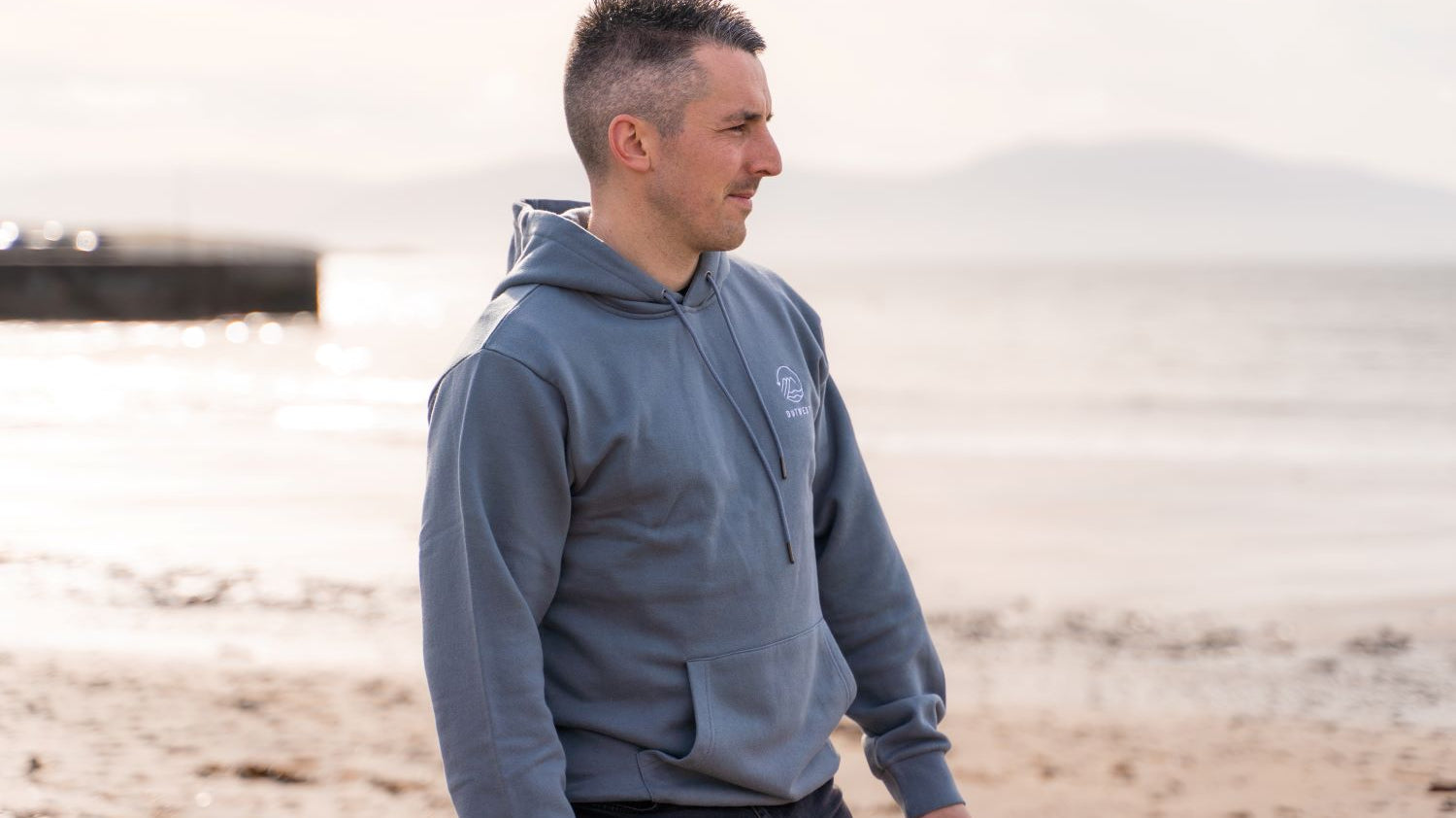 Man on beach looking out to sea, wearing blue Outwest Hoodie