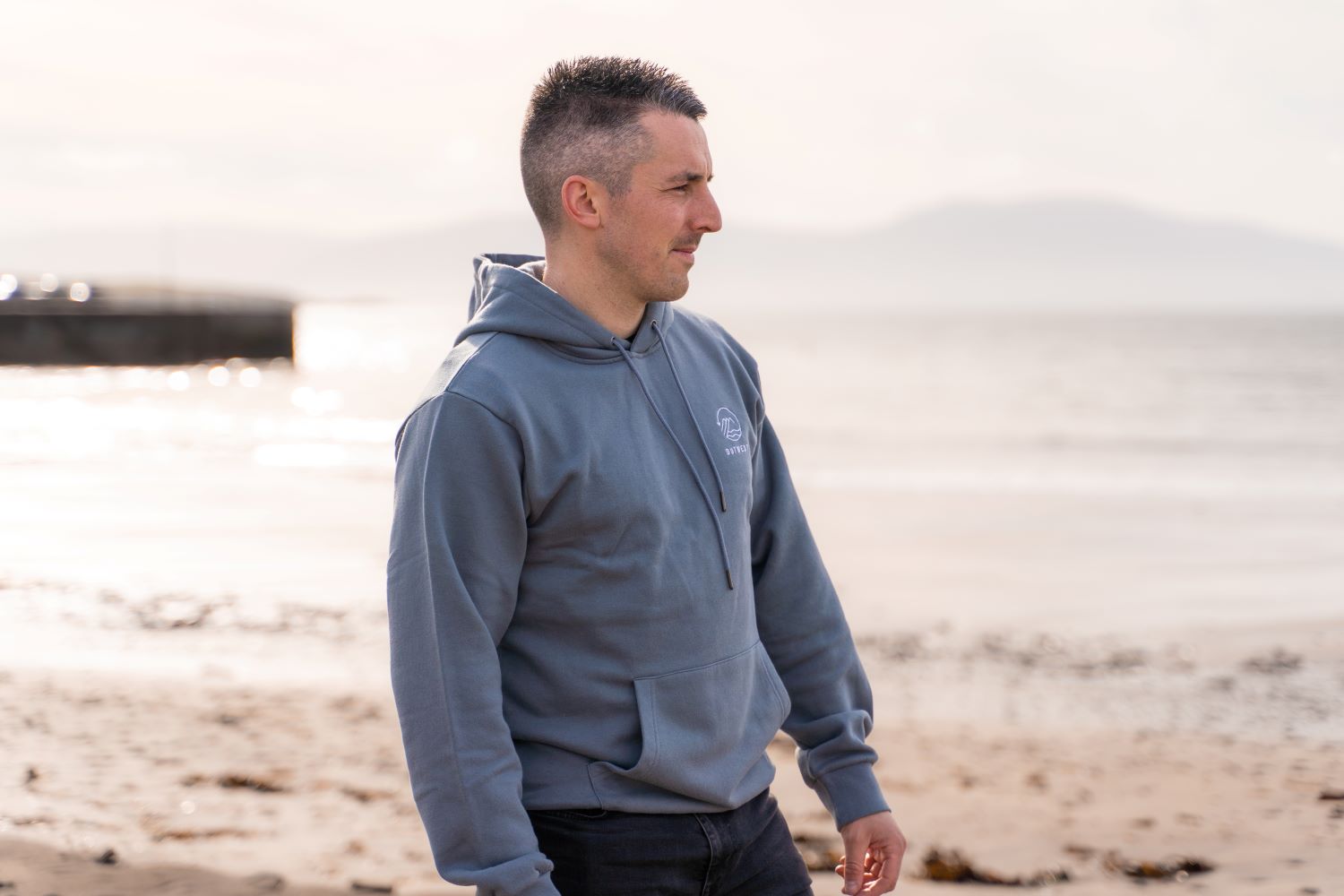 Man on beach looking out to sea, wearing blue Outwest Hoodie